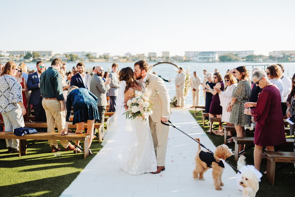 Newlyweds celebrate at outdoor waterfront ceremony with dog joining them on white aisle runner.