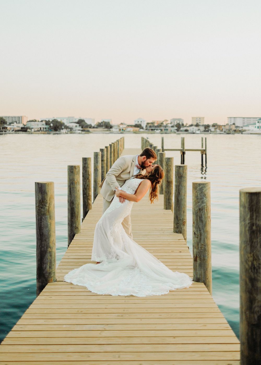 A bride wearing a flowing white dress poses gracefully on a wooden dock overlooking coastal waters during sunset.