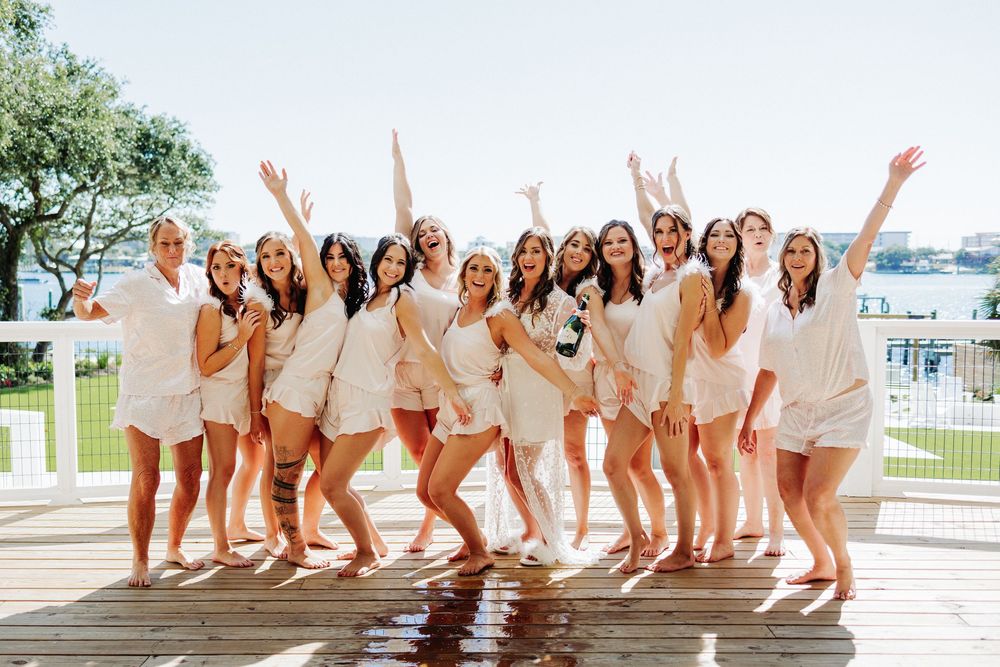 A group in coordinated white and beige outfits poses excitedly on a waterfront deck during a sunny day.