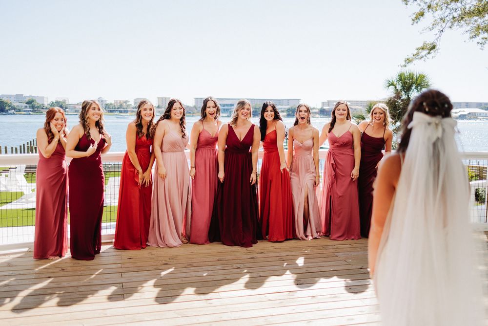 Bridesmaids in alternating red and blush colored dresses pose together with the bride on a sunny outdoor patio.