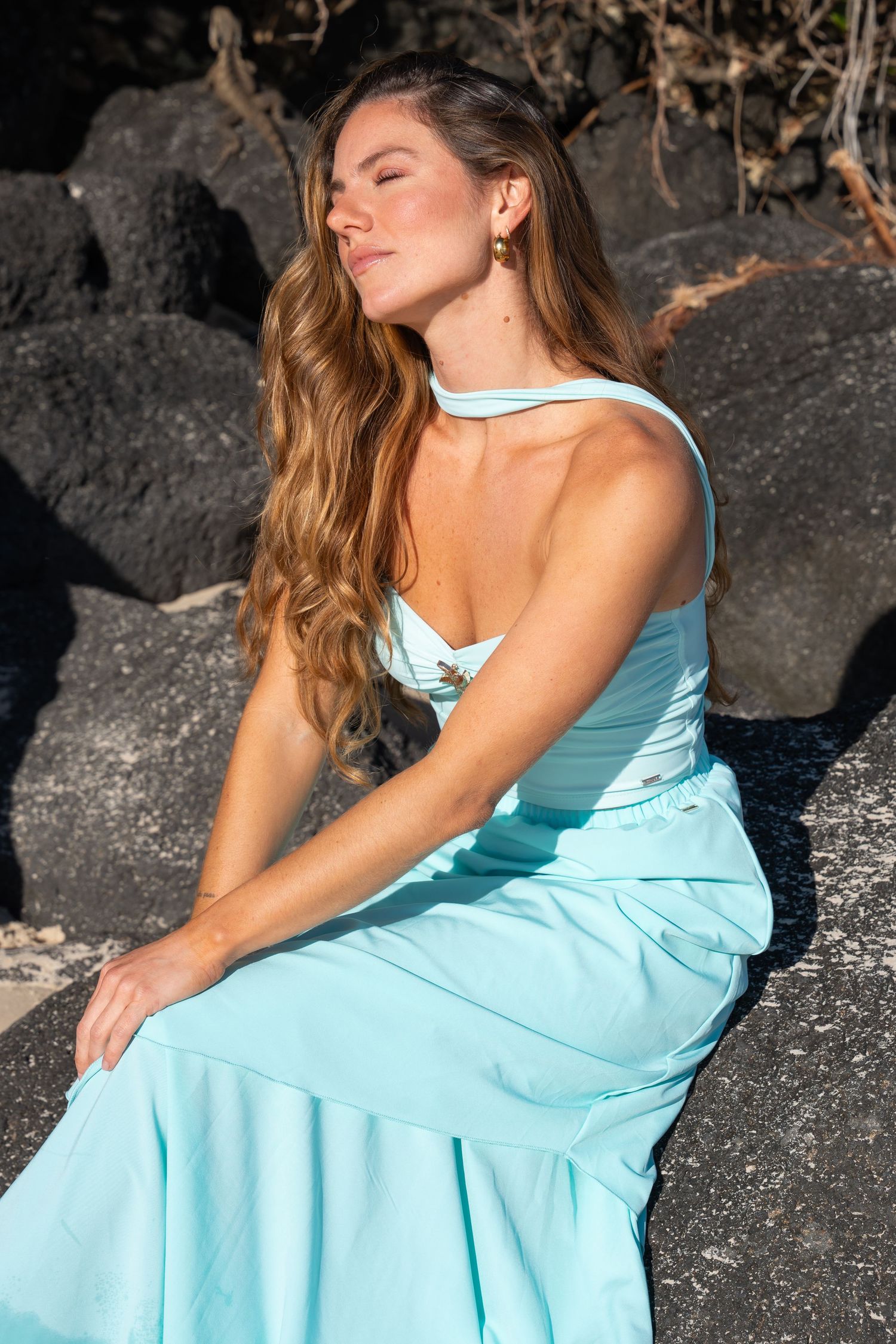 A fashion model in a light blue sundress poses serenely on black volcanic rocks in sunlight in Coolangatta.
