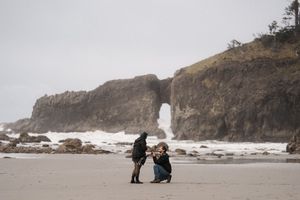 Two people share a special moment on a foggy beach with dramatic coastal cliffs and natural arch in the background.