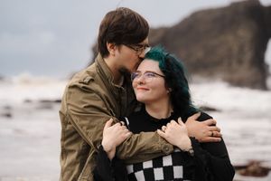 A romantic couple in brown jackets embrace near a windy beach on a cloudy winter day.