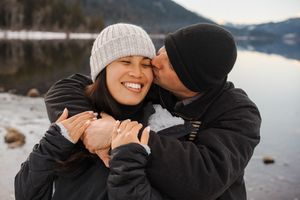 Romantic winter engagement photography session by a lake with cozy sweaters and beanies in scenic mountain setting.