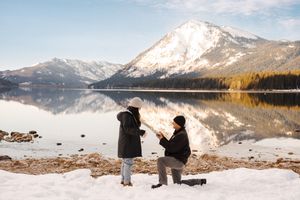 Two people kneel at a snowy mountain lake shore with a stunning snow-capped peak reflected in the calm water.