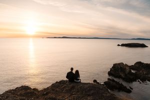 Couple sitting on rocky cliff watching golden sunset over calm ocean waters with distant islands on the horizon.
