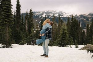Two people embrace while standing in snowy mountains surrounded by evergreen trees and mountain peaks in the background.