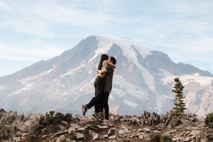 Two silhouettes embrace on a rocky overlook with snow-capped Mount Rainier rising majestically in the background.