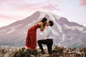 A couple in red and black clothing embraces against a scenic mountain backdrop at sunset.