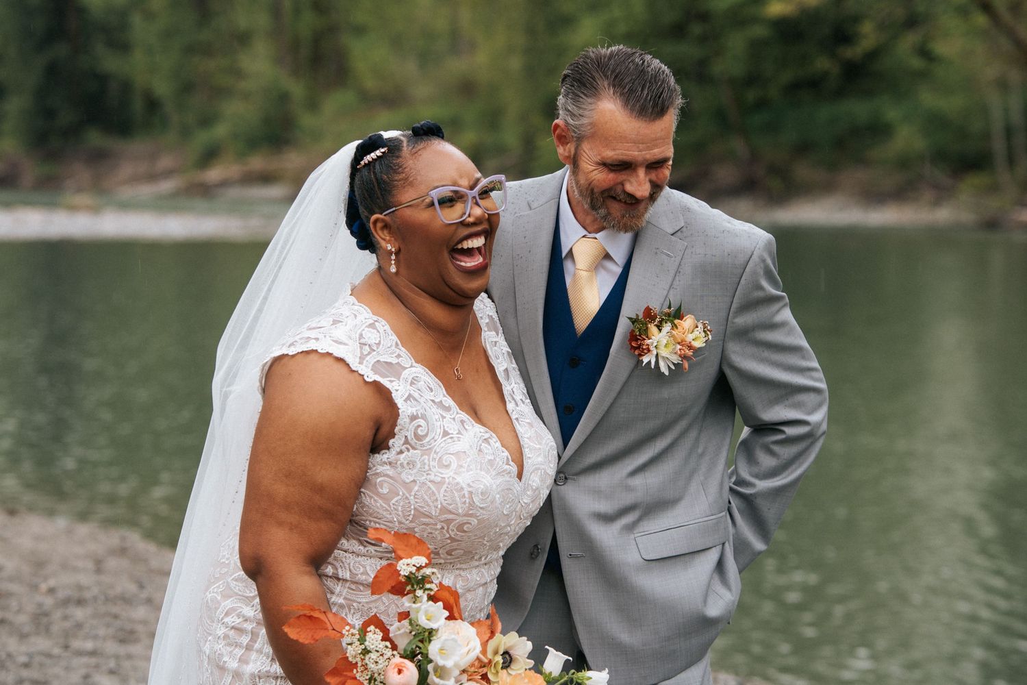 A joyful wedding couple stands together by a serene lake, the bride in white lace and groom in a grey suit.