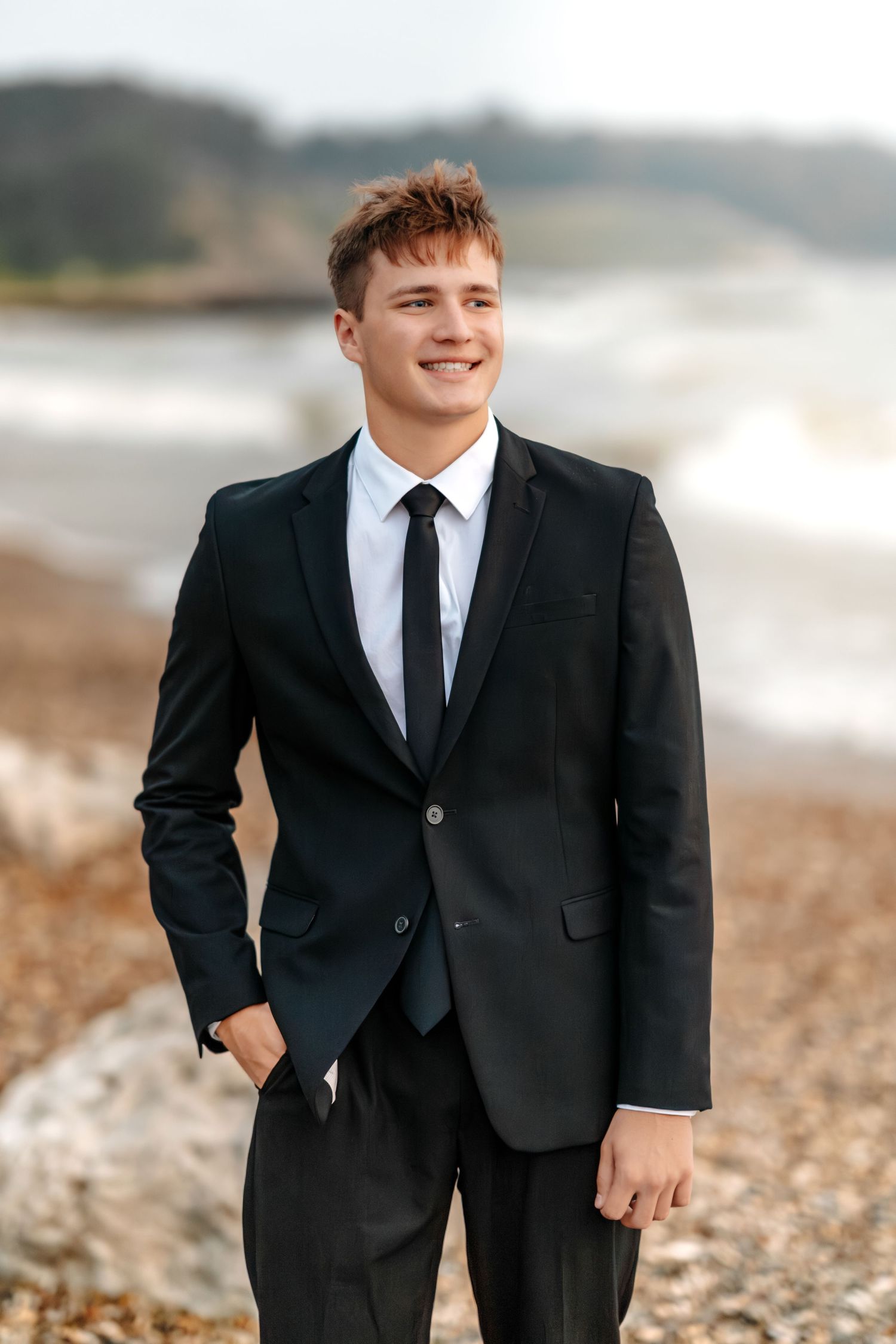Well-dressed person in black suit and tie smiling while standing on rocky beach with ocean waves in background.