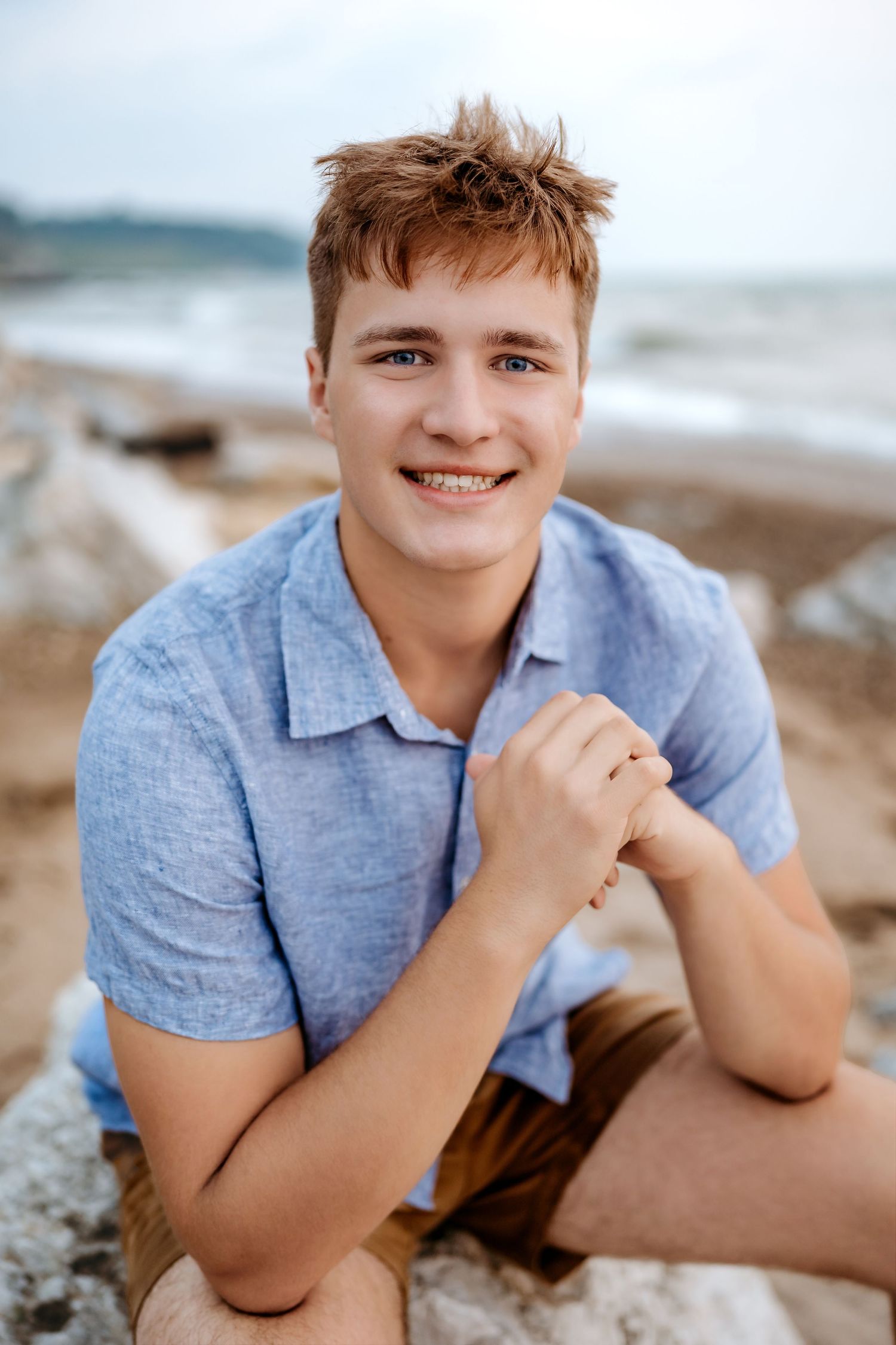 Someone in casual attire poses on a rocky beach with a natural smile and windswept hair.