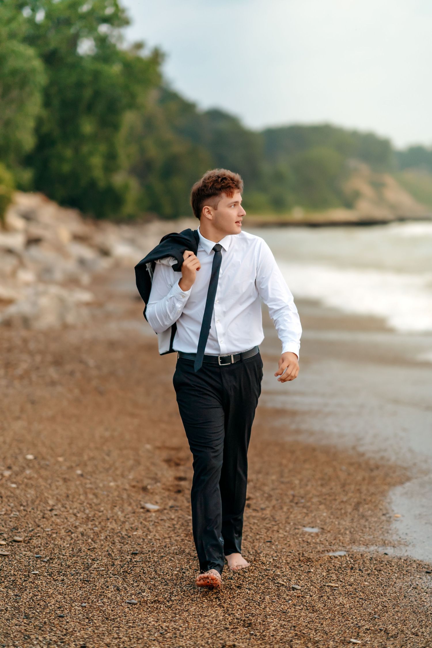 A person in business attire walks barefoot along a rocky beach while carrying a dark jacket over their shoulder.