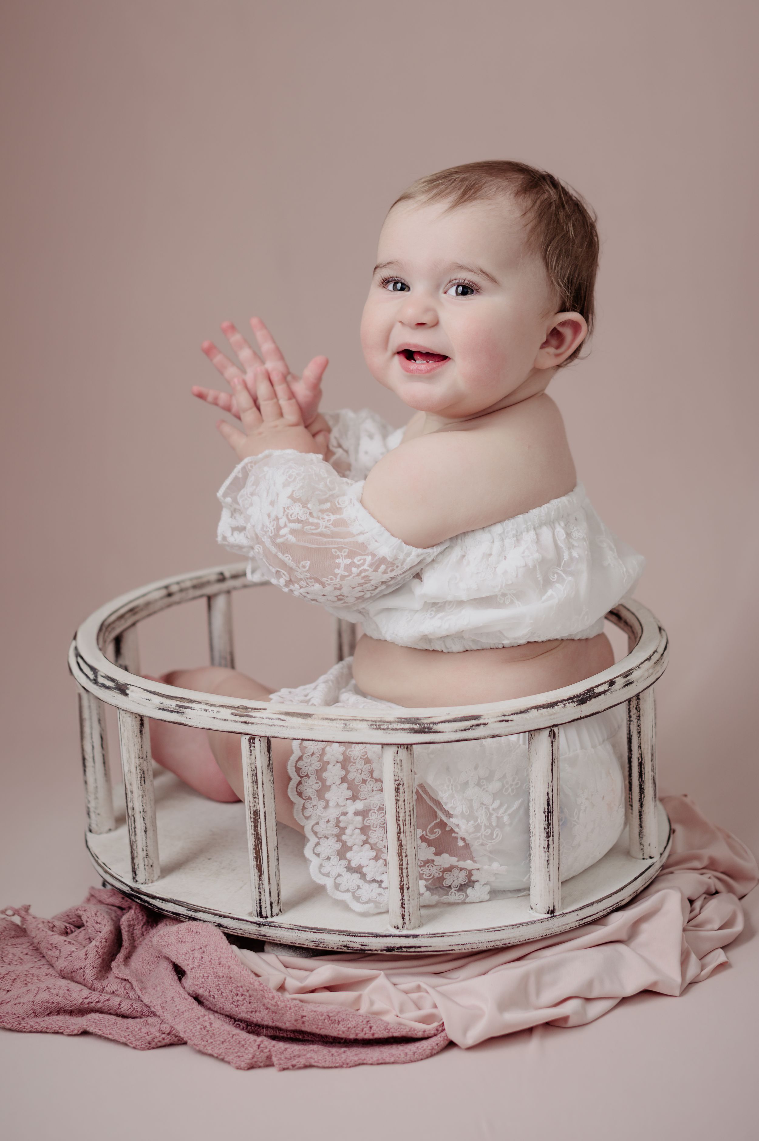 A smiling baby wearing white lace sits in a vintage-style white circular prop during a portrait session.