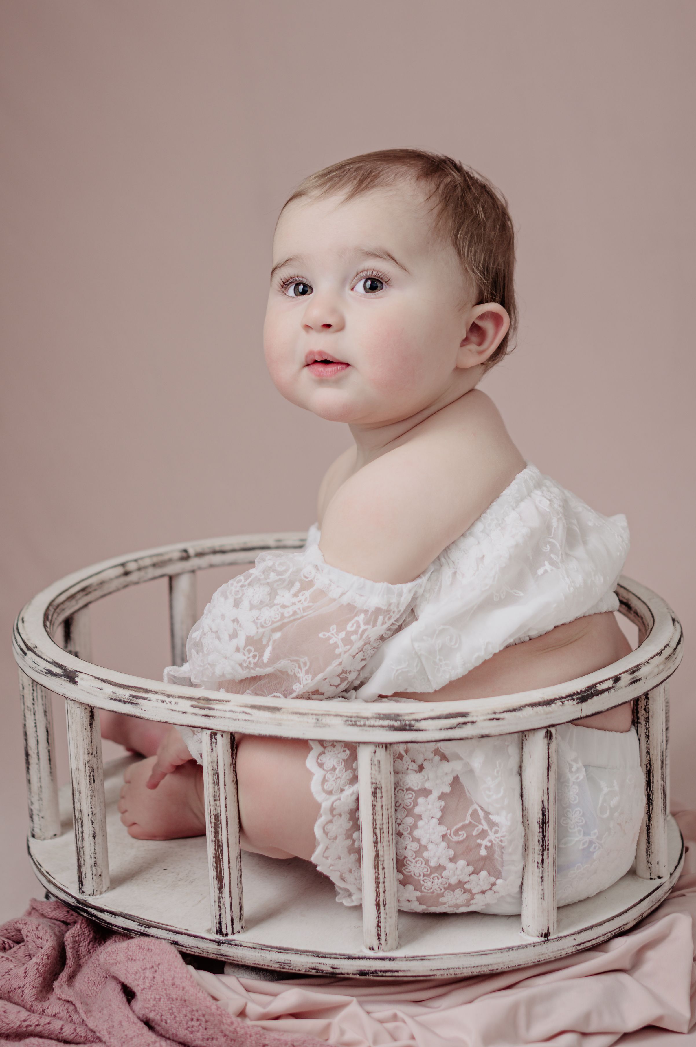 A baby in a white lace dress sits inside a distressed white circular wooden prop against a pink backdrop.