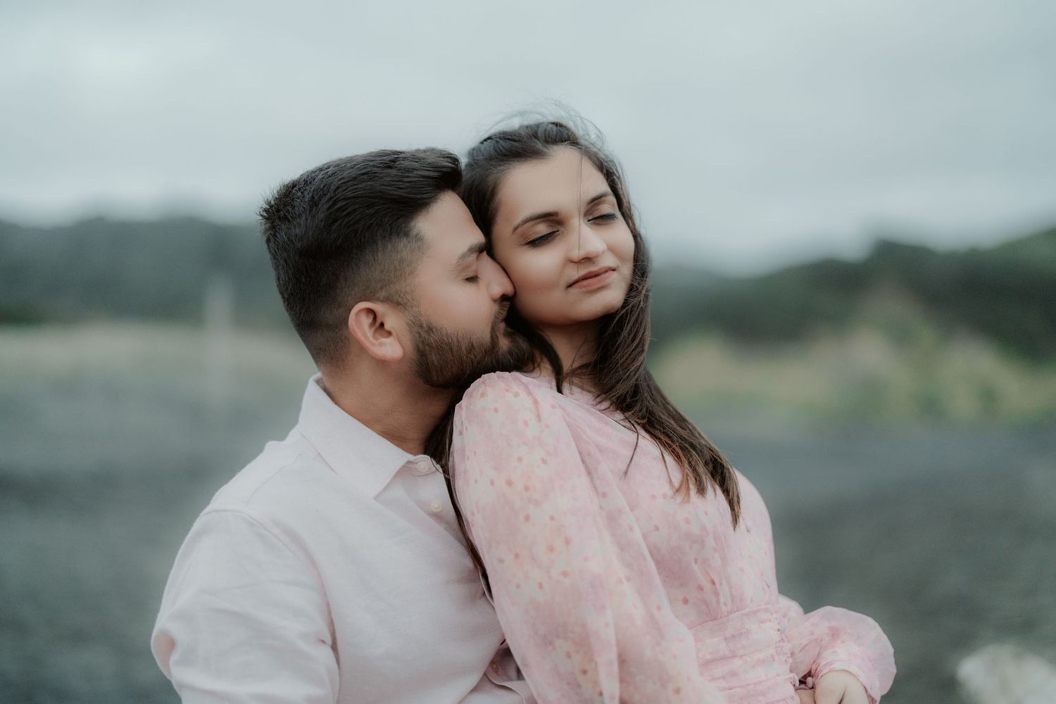 A romantic couple in matching pink outfits share an intimate moment outdoors against a blurred natural background.
