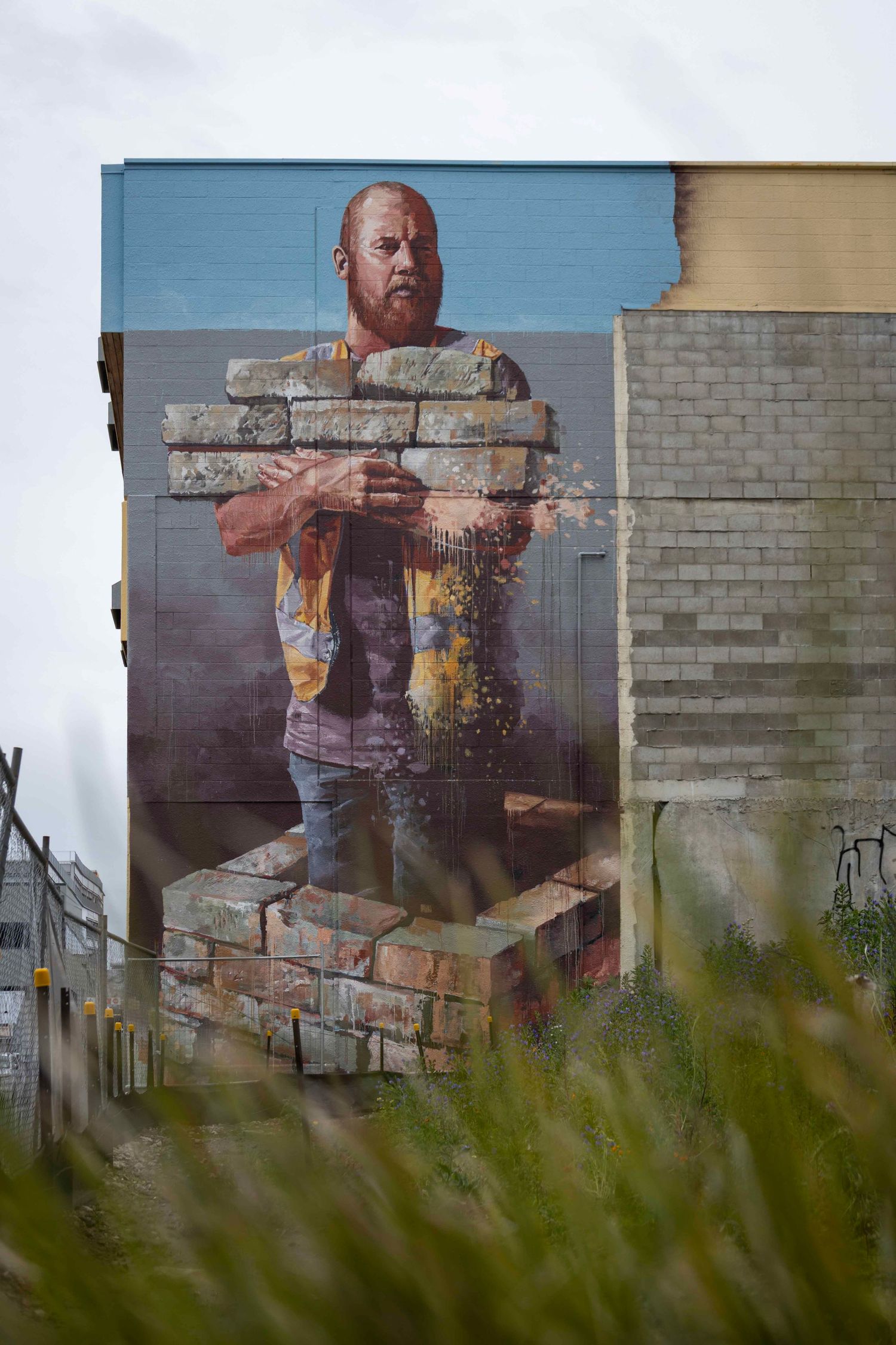 Street mural of a builder holding disappearing bricks, painted on a concrete wall with wild grass in the foreground.