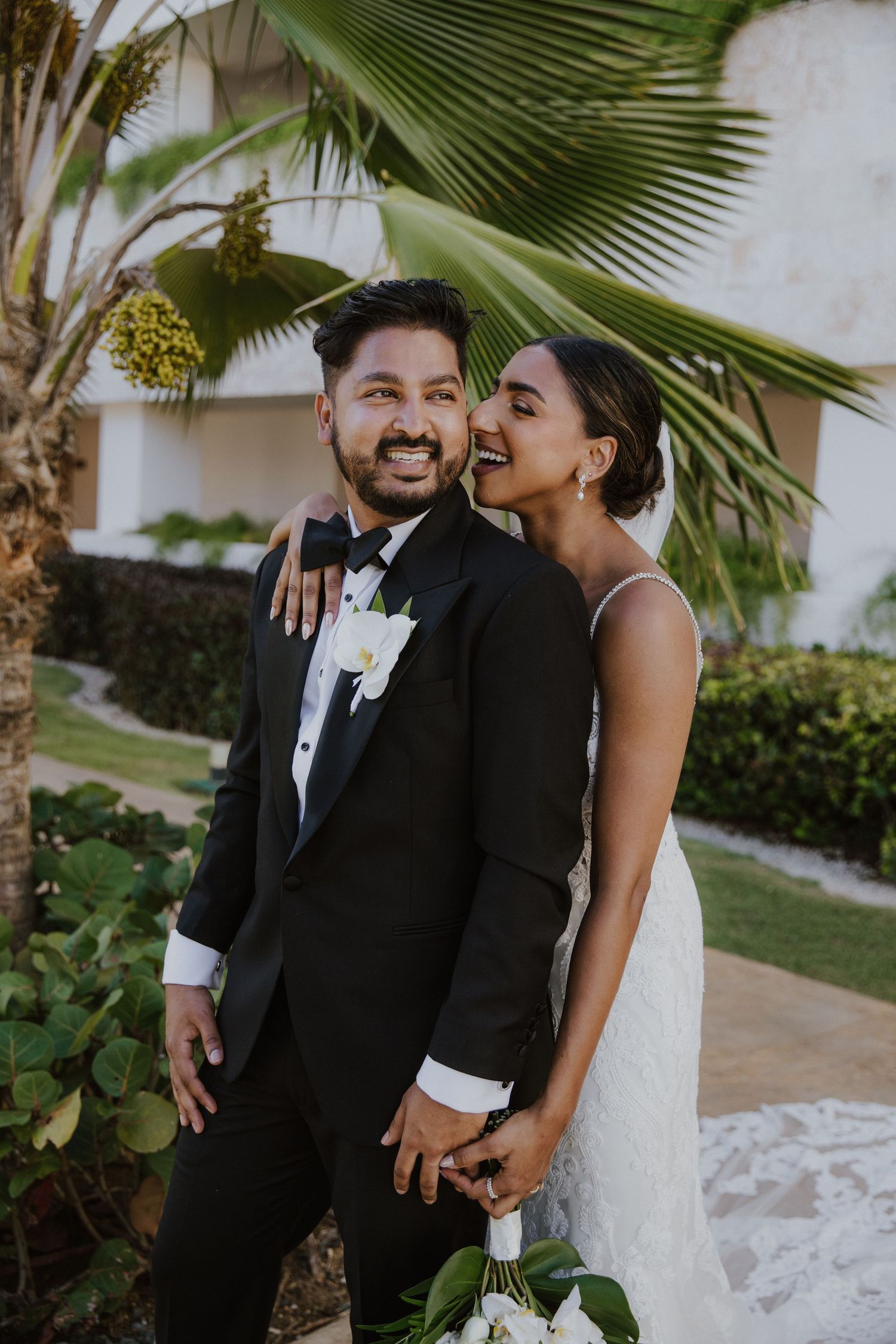 Joyful wedding couple sharing a laugh among palm trees in a tropical setting.