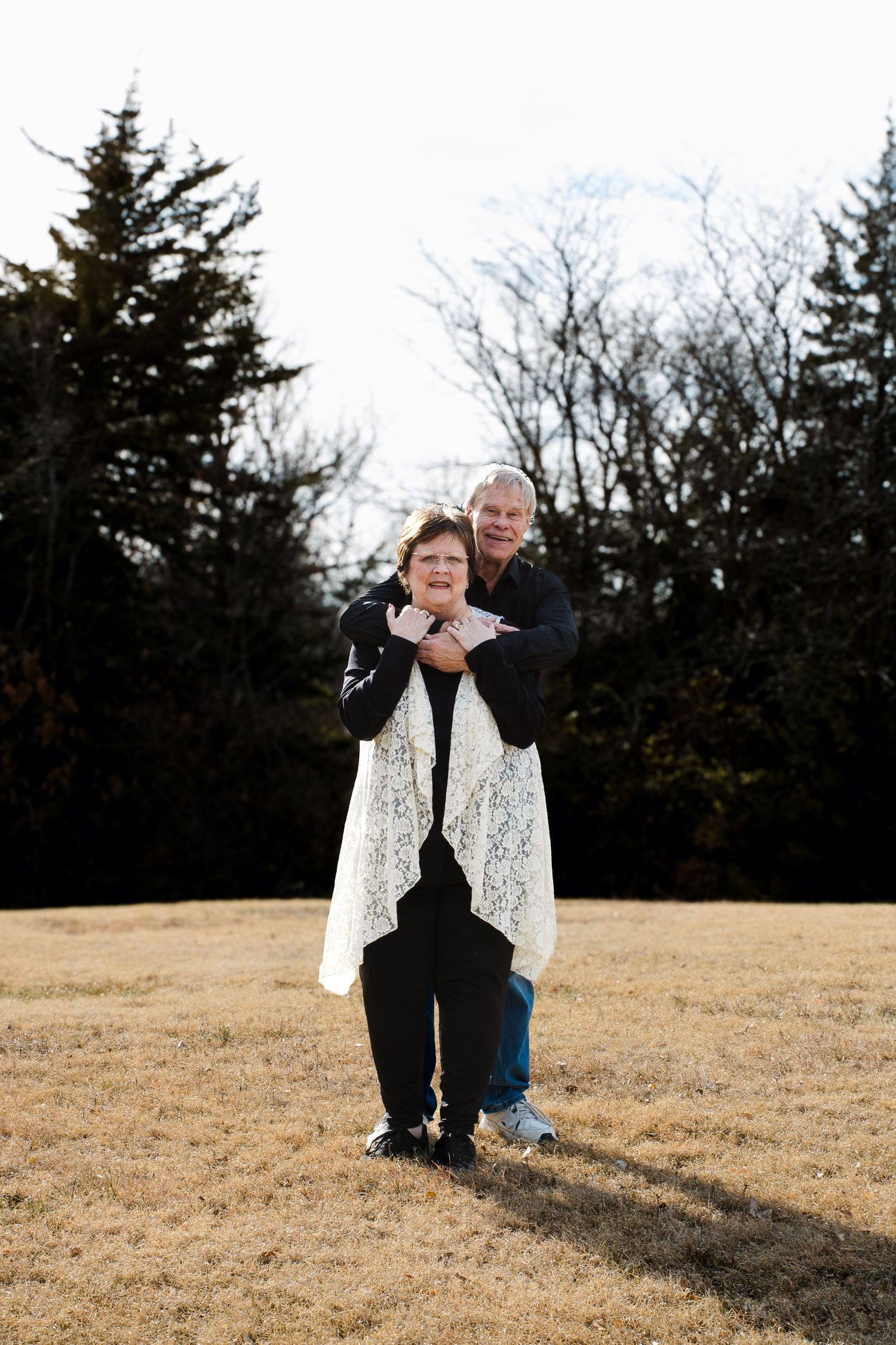 A couple stands together in a field with evergreen trees in the background during winter.