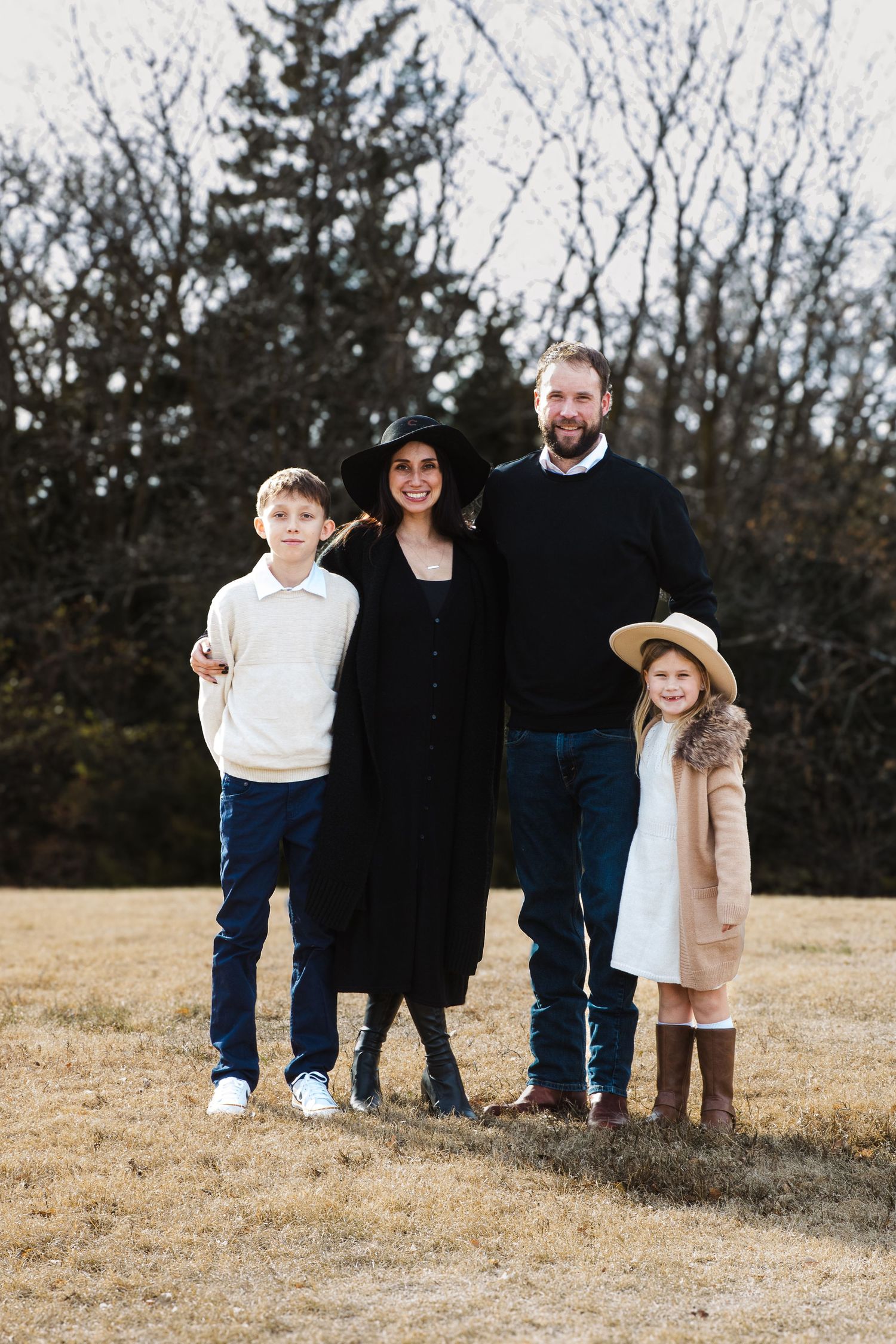 A family stands together outdoors in winter clothing against a backdrop of bare trees and evergreens.