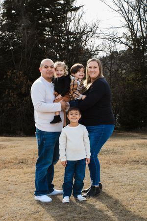 A family of five poses together outdoors in casual clothing on a grassy field with trees in the background in Kearney, NE.