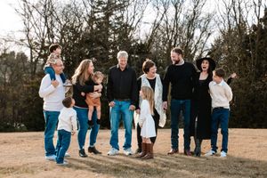 Extended family group wearing casual blue jeans and sweaters stands together outdoors in a natural field setting during golden hour.