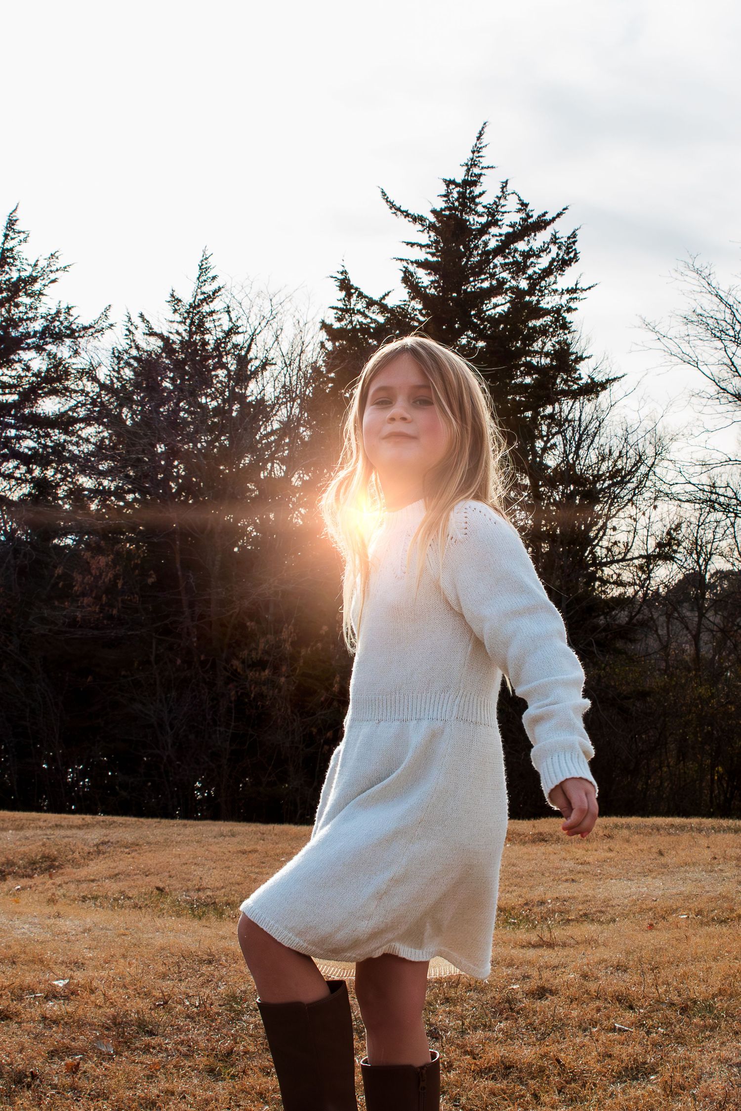 A girl in a white sweater silhouetted against golden sunlight in a rural field with evergreen trees and dry grass in Kearney, Nebraska.