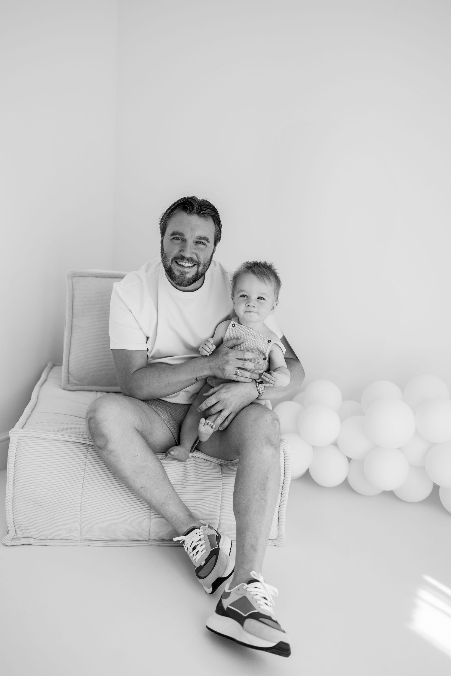 Black and white portrait of an adult and young child sitting together in a white chair with balloons visible in background.