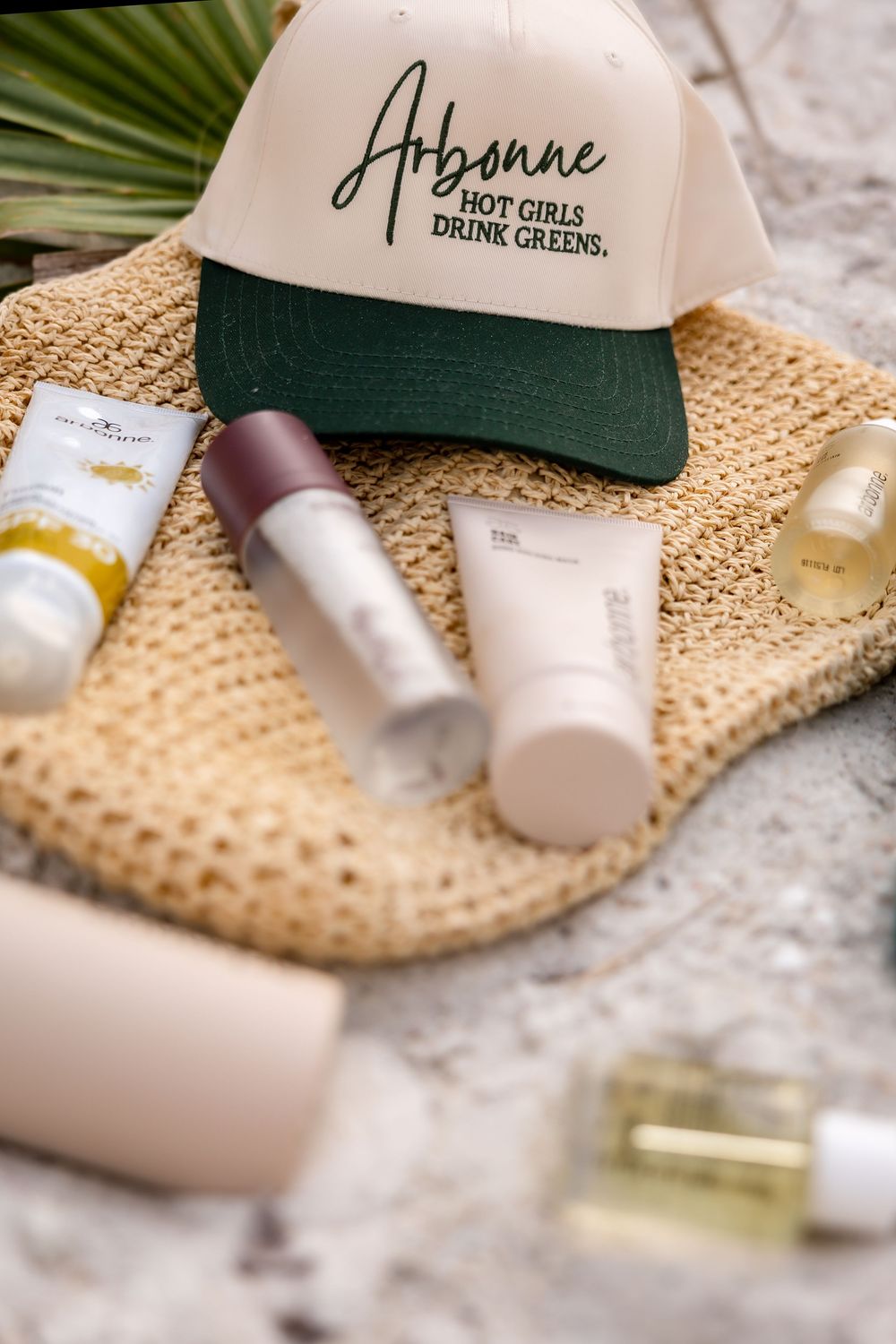 Arbonne Hot Girls Drink Greens cap displayed on woven straw bag surrounded by Arbonne beauty and skincare products.