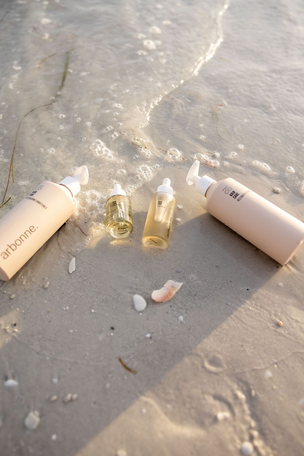 Arbonne skincare bottles and serums arranged on wet sandy beach shore with gentle ocean waves in golden sunlight.