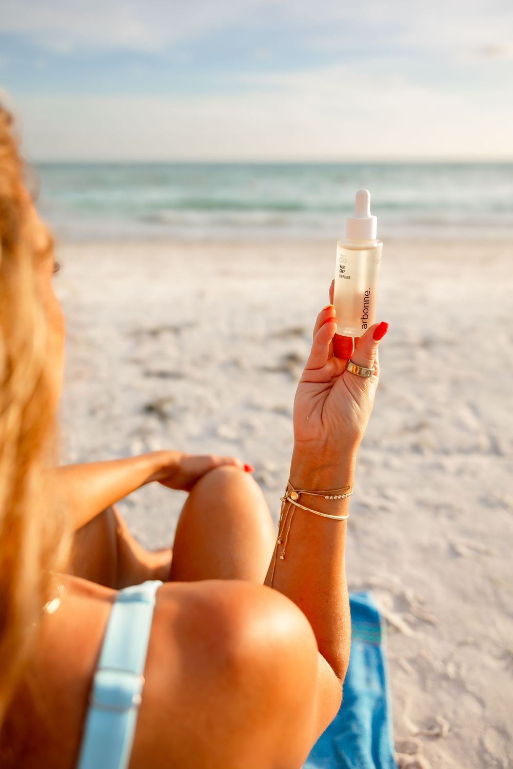Woman in light blue bikini holds up Arbonne serum bottle while sitting on a sunny beach with ocean in background.