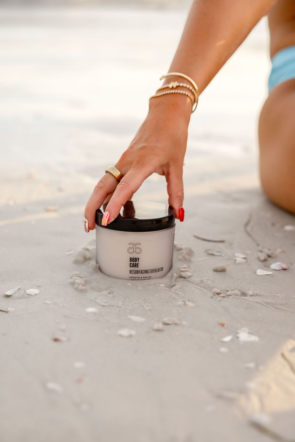 Tanned woman with red nails and gold bracelets reaches for Arbonne RE9 Body Care rejuvenating cream on sandy beach.