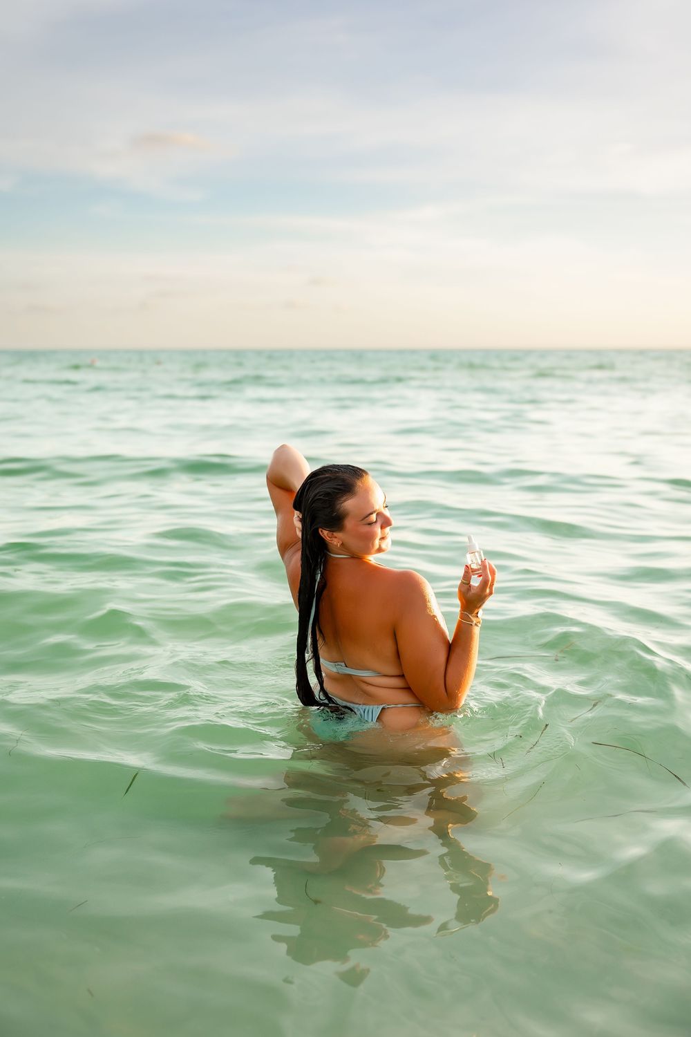 Woman in bikini standing in calm turquoise ocean water at sunset holding a drink.