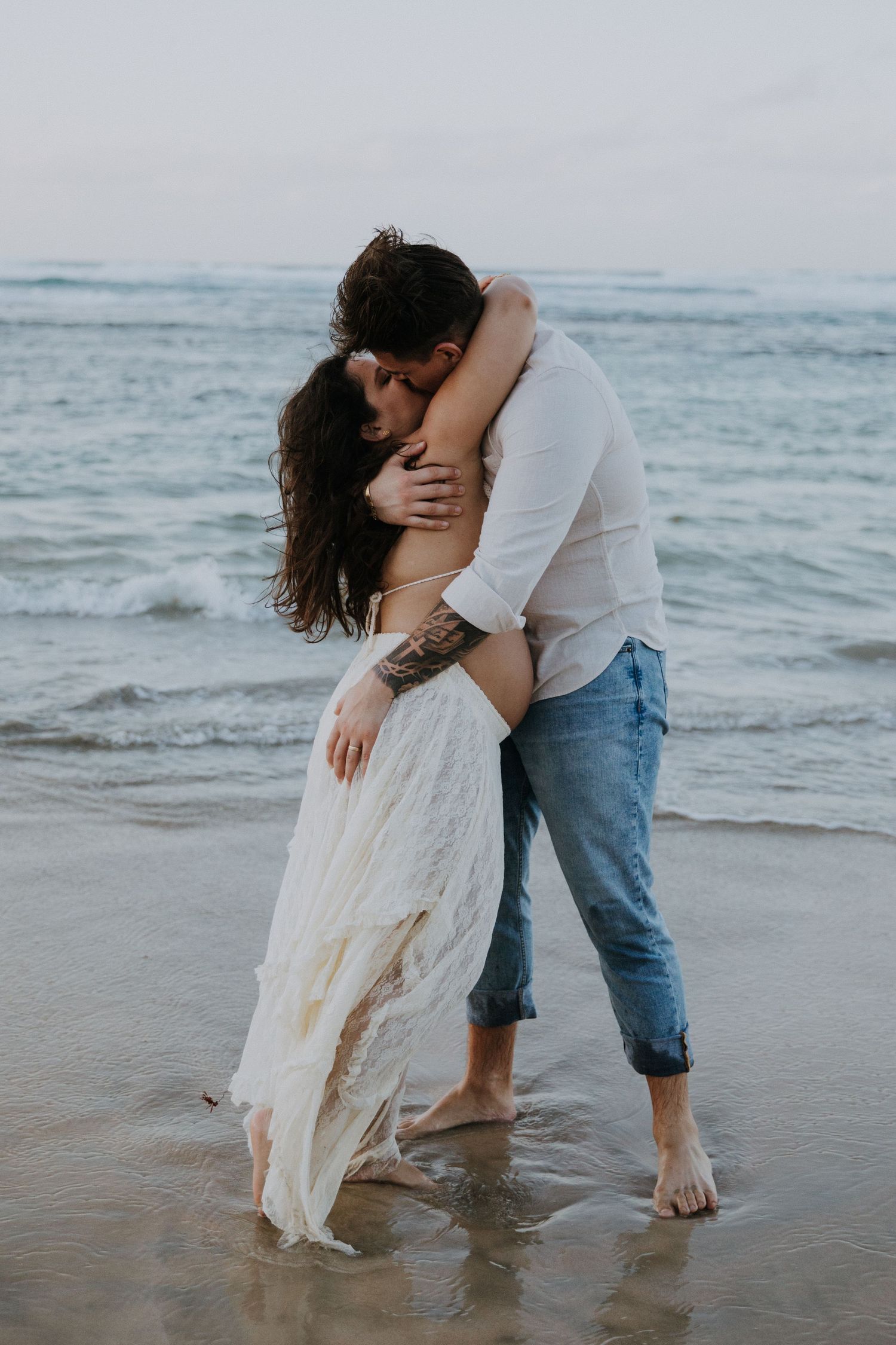 Romantic couple embrace on a beach at sunset with waves in the background.
