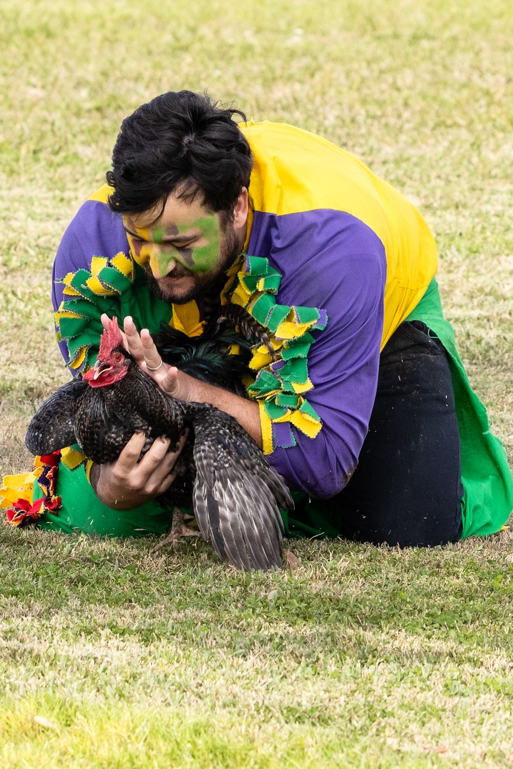 Person in colorful Mardi Gras costume playing with chicken on grassy field.