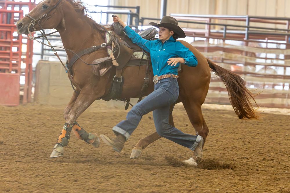 A rodeo competitor dismounts from a running horse during a steer wrestling event in a dirt arena.