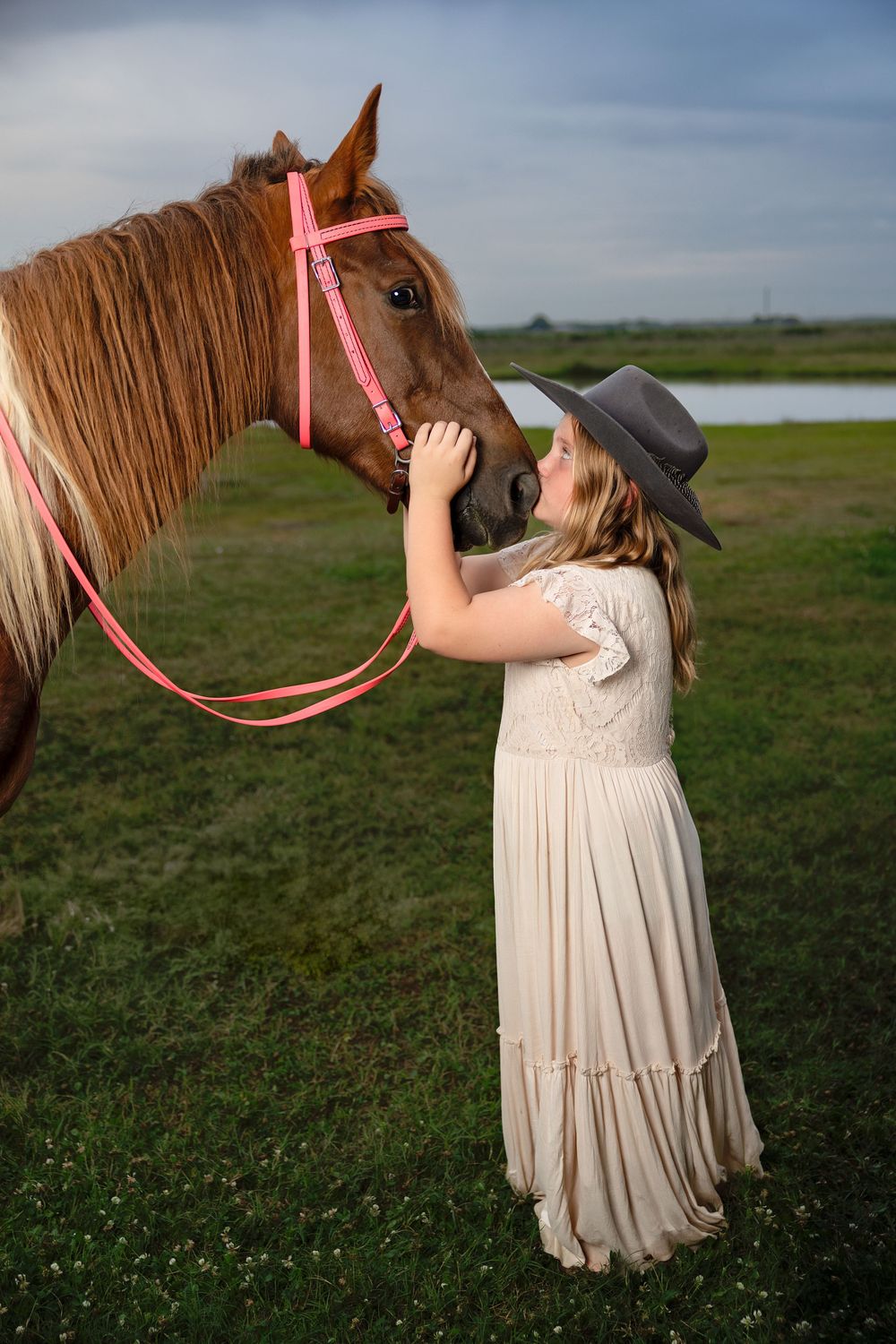 Girl in cream dress and gray hat kissing a chestnut horse with pink halter in a green field.