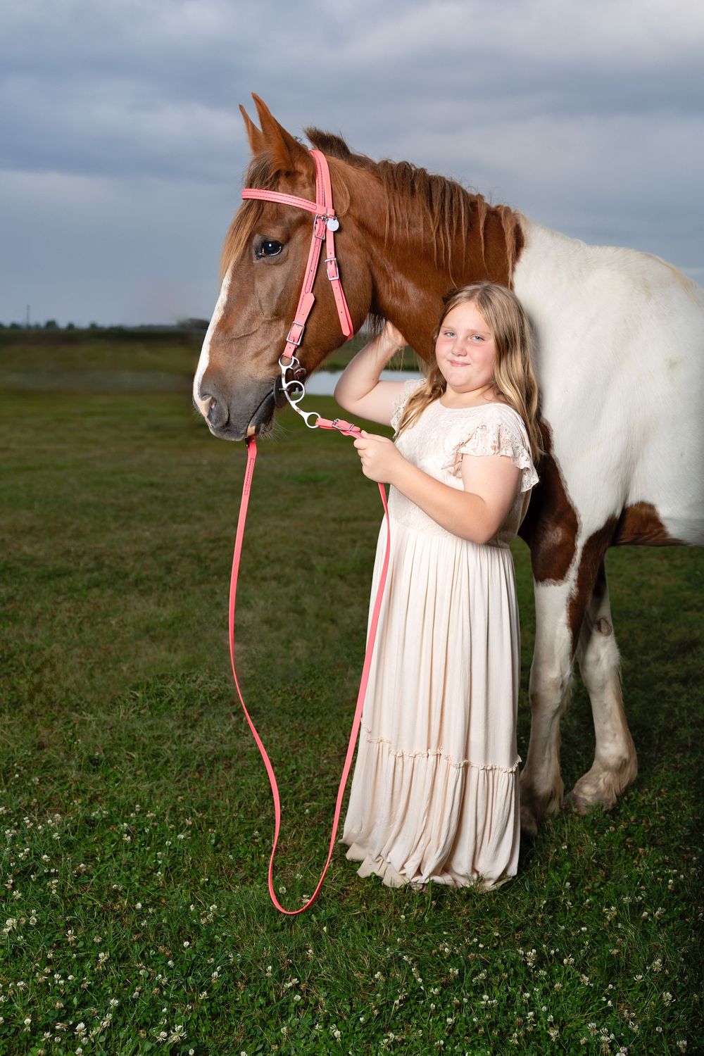 A young girl in a white dress holds the pink lead rope of a chestnut and white paint horse in a green field.