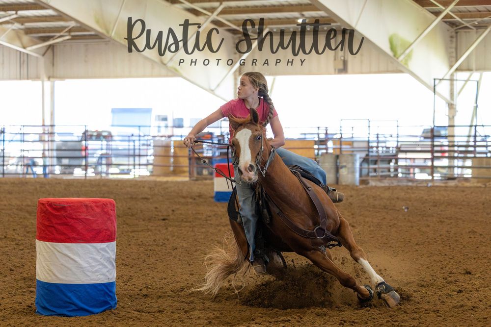 Young cowgirl on horseback performing barrel racing around a red, white, and blue barrel in an indoor rodeo arena.