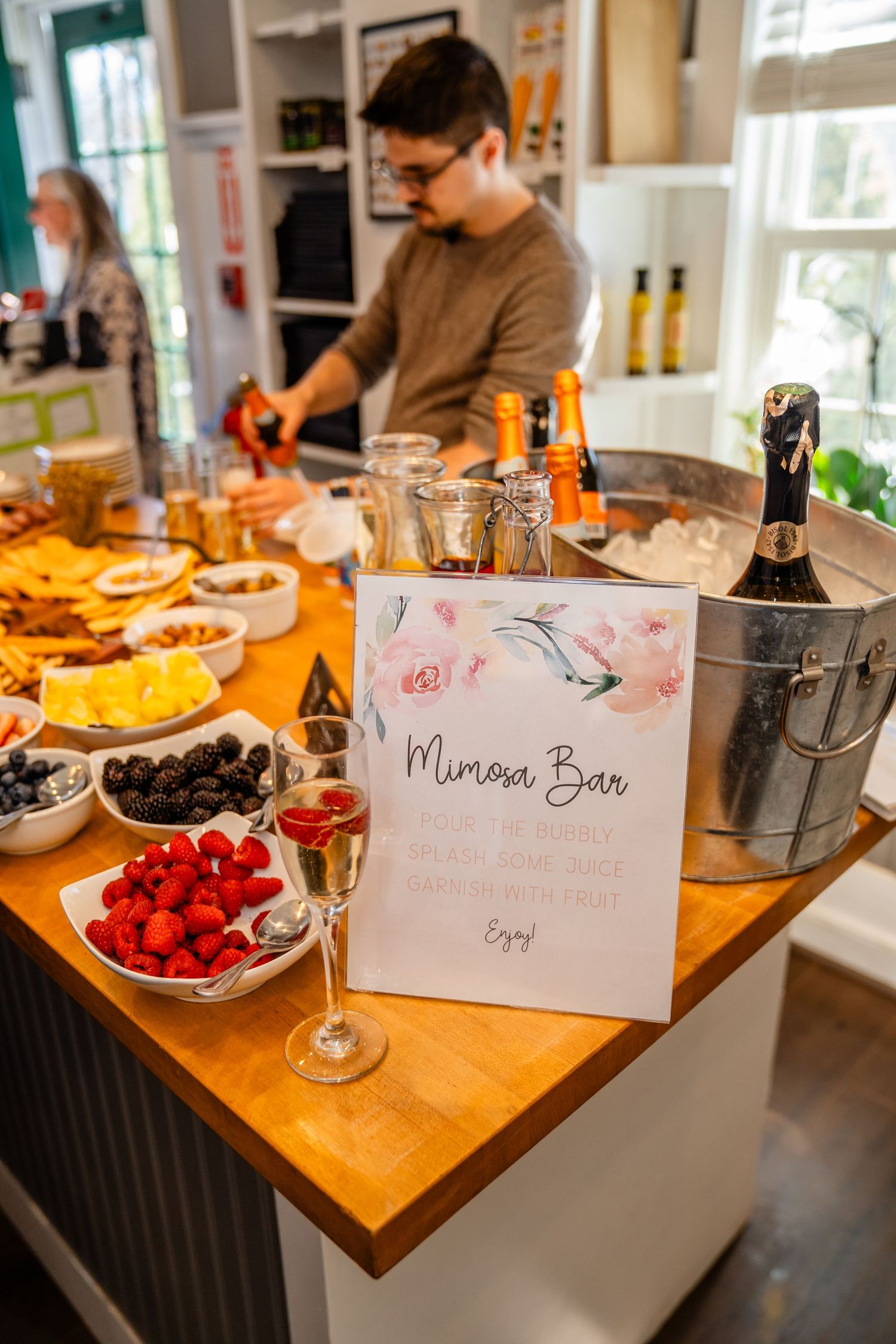 A mimosa bar setup with fresh fruit, champagne in an ice bucket, and a decorative sign on a wooden counter.