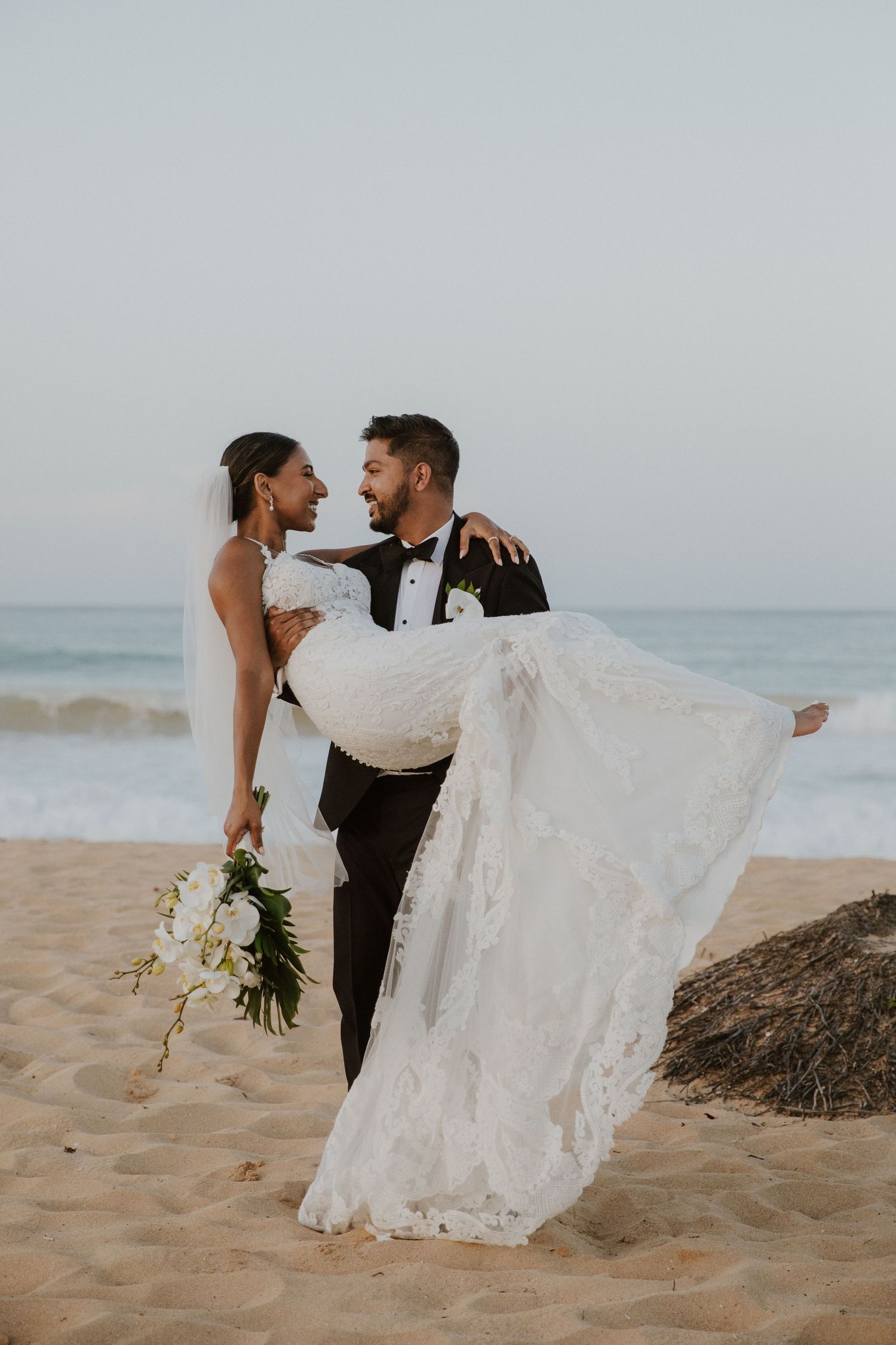 Groom carrying bride in lace dress on beach with rocks and ocean waves in background.