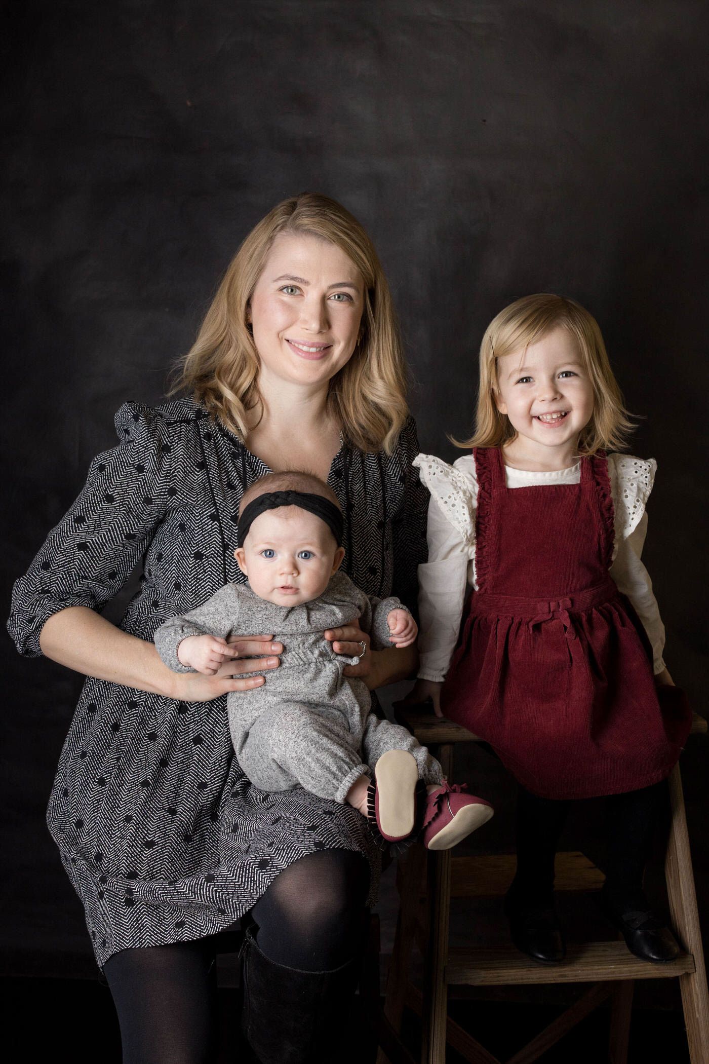 Studio portrait of a family against dark background, with one child in red dress and a baby in grey outfit.