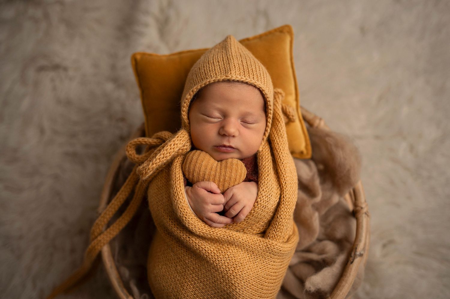 Sleeping swaddled newborn baby wrapped in soft golden knit material against a textured beige backdrop.
