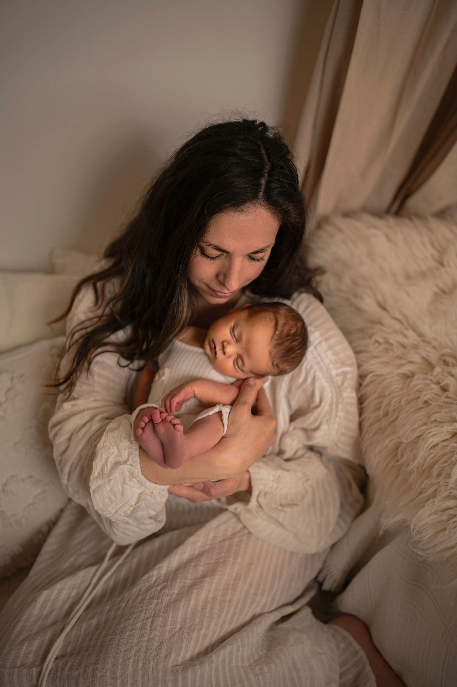 A mother in a white sweater lovingly holds her sleeping newborn baby on a cozy bed with soft lighting.