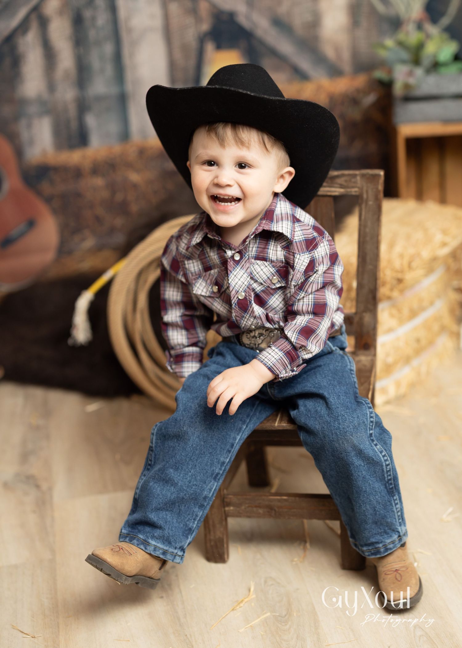 Young child in a cowboy hat and plaid shirt sits on wooden chair in rustic farm setting.