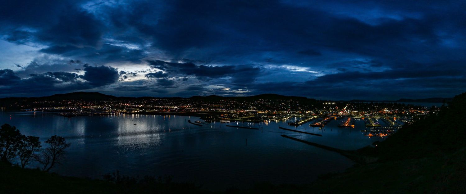 Dramatic nighttime cityscape panorama reflected in calm waters under dark blue sky.