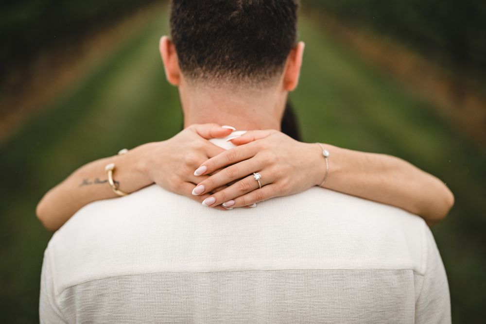 Couple in white outfits embracing from behind in a romantic outdoor setting.