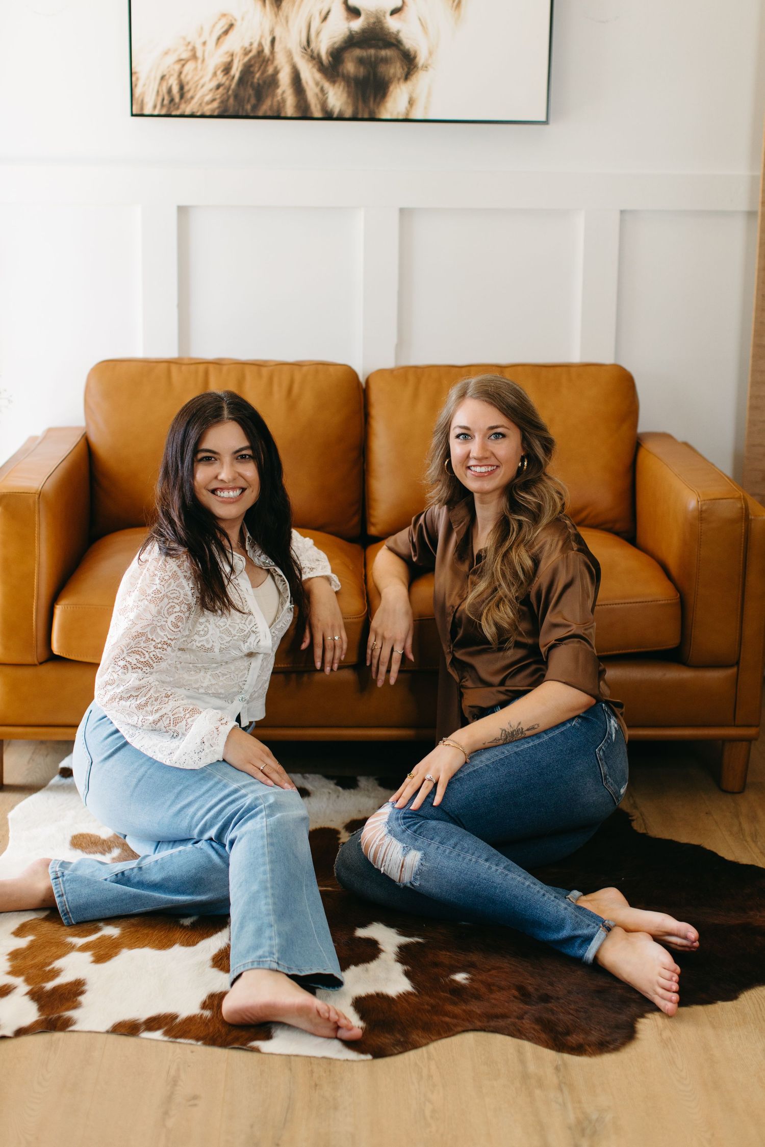 Two people relaxing on a tan leather couch with a cowhide rug on hardwood floors beneath an animal wall print.