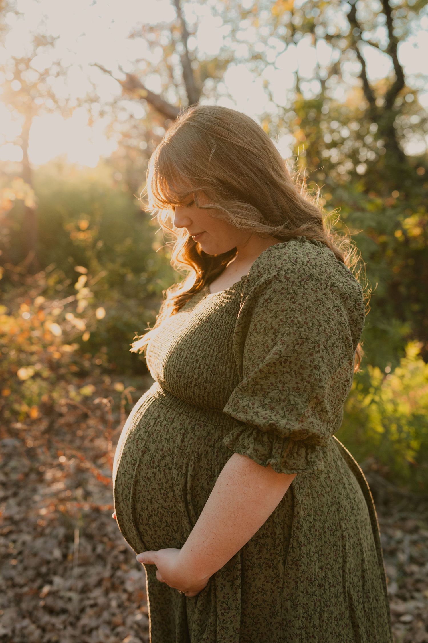 Silhouetted pregnant figure in a green dress embraces belly during golden hour maternity photoshoot in nature.