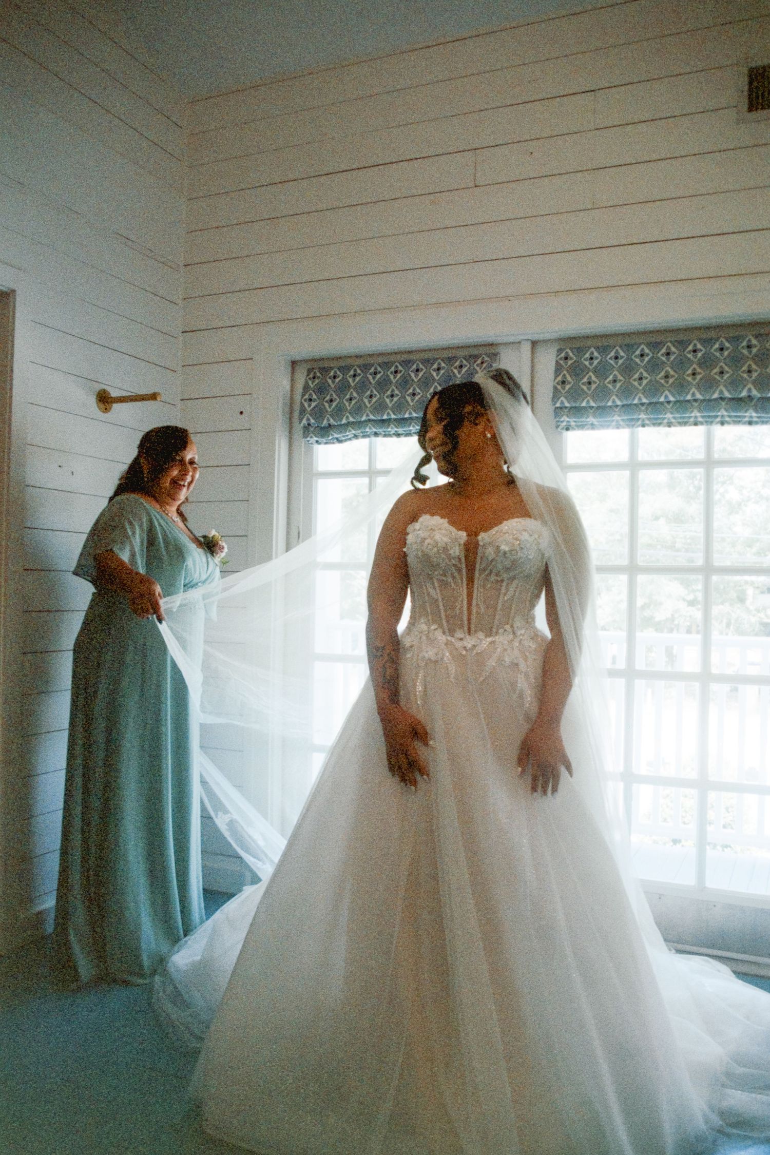A white wedding gown with delicate lace details, tulle skirt, and off-shoulder design is showcased in a bright sunlit room.